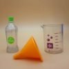 A clear plastic vinegar bottle with painted green bottle cap and label, a yellow plastic funnel, and a clear plastic beaker with purple measuring decals.